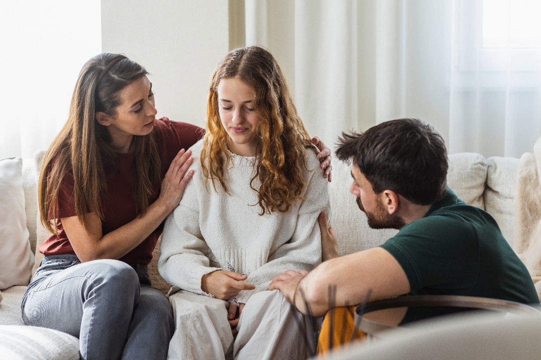 Teen girl being comforted by her parents as they sit on the couch.